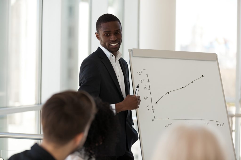 Workers sitting at modern conference room in office listening black team leader, focus on african experienced coach teaches company staff make presentation about increasing productivity and revenue